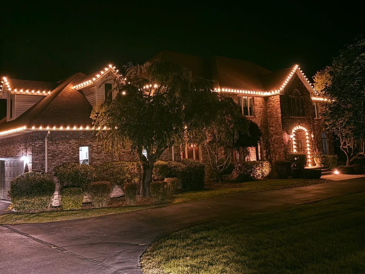 Brick two-story home at night with warm white holiday lights along the rooflines, peaks and entry arch